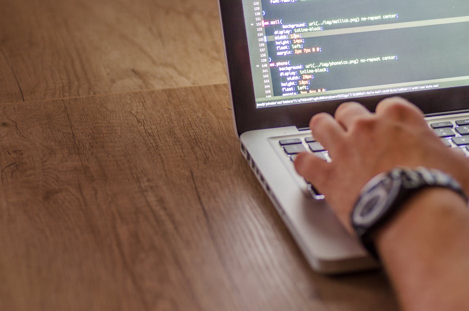 This image shows a person viewing a computer monitor in an office environment during daytime. The screen displays lines of code in multiple colors, indicating programming or software development work.