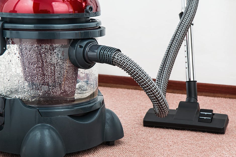 Close-up of a modern vacuum cleaner with a transparent dust container filled with debris and a flexible hose attached, positioned on a plush, pink carpet in a residential setting. The vacuum cleaner f